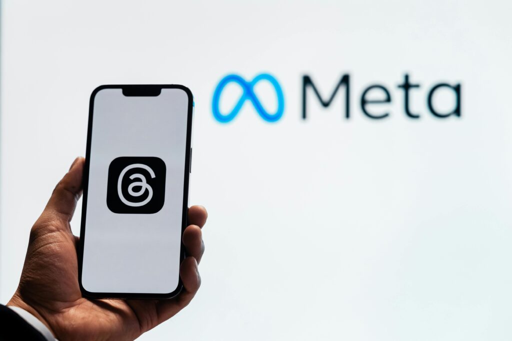 What is Meta Adds A hand holding a smartphone showing the Threads app with Meta logo in the background.
